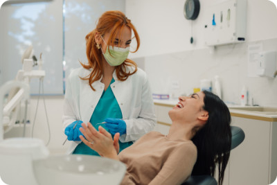 Weiblicher Zahnarzt mit chirurgischer Maske, die zahnaerztliche Werkzeuge haelt und mit gluecklicher Patientin spricht, die im Zahnarztstuhl in einer hellen modernen Zahnklinik sitzt.